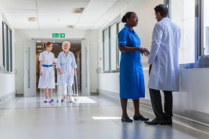 A nurse and doctor talk in a hospital corridor whilst another staff member walks besides a patient